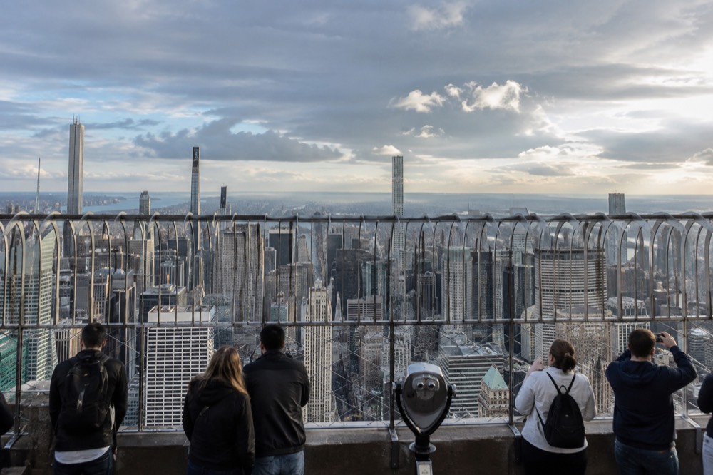 Visitors standing along the open-air railing of the Empire State Building observation deck overlooking Manhattan