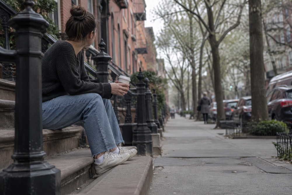 A person sitting alone on a stoop of a Brooklyn brownstone, resting with a coffee on a quiet morning street