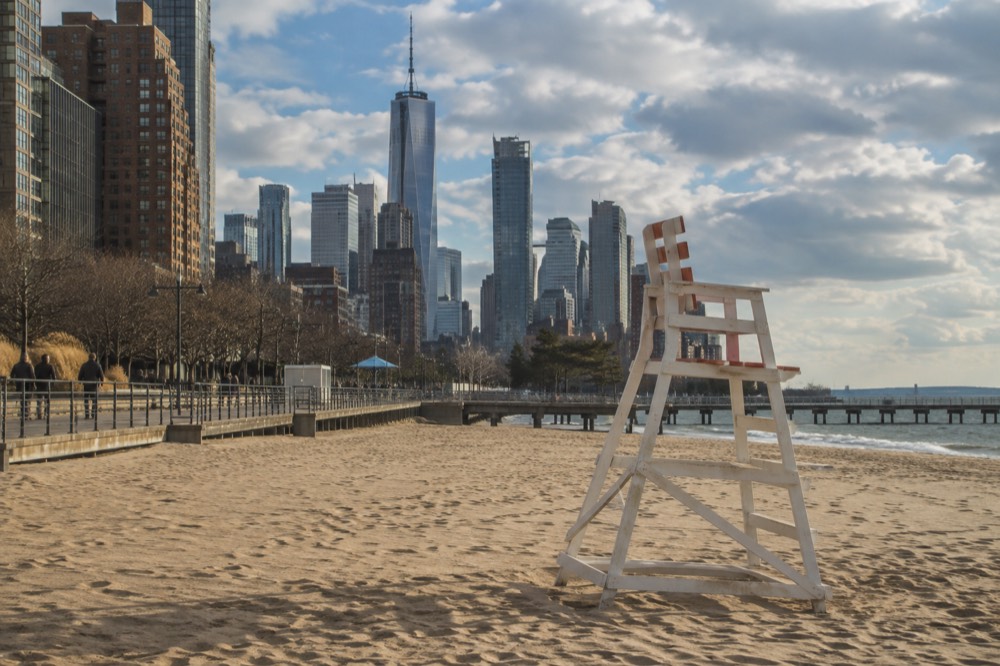 A lifeguard chair standing on a wide sandy beach with a boardwalk and city skyline behind it.