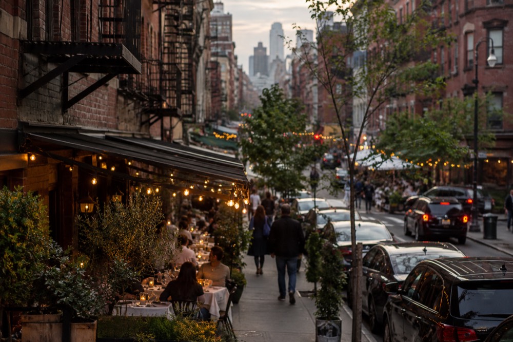 Restaurant tables and chairs set out on a narrow sidewalk under string lights in the evening