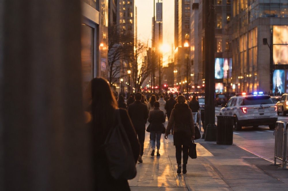 Evening crowds walking along a brightly lit Manhattan avenue with a police patrol visible on the corner