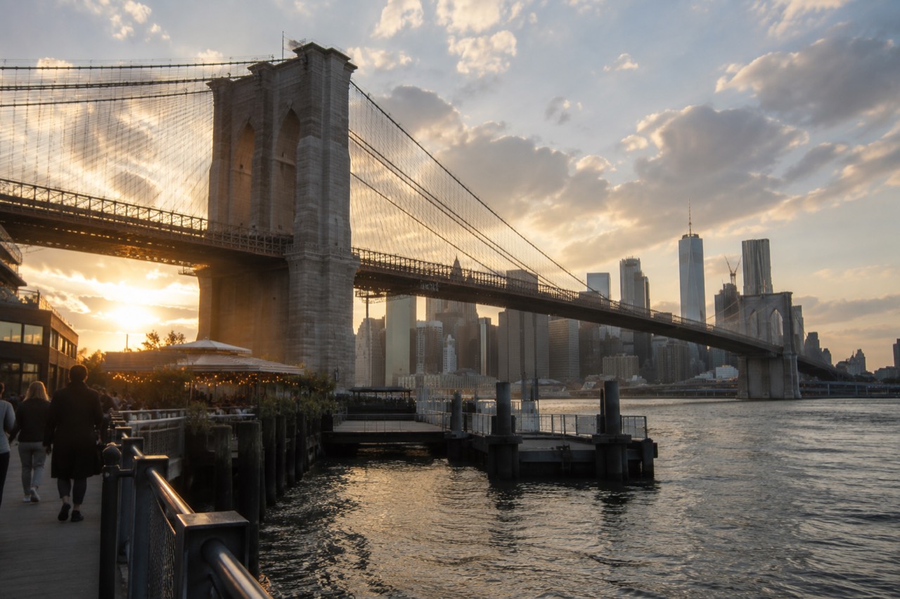 The Brooklyn Bridge rising over the waterfront restaurants and piers on the Brooklyn side at golden hour