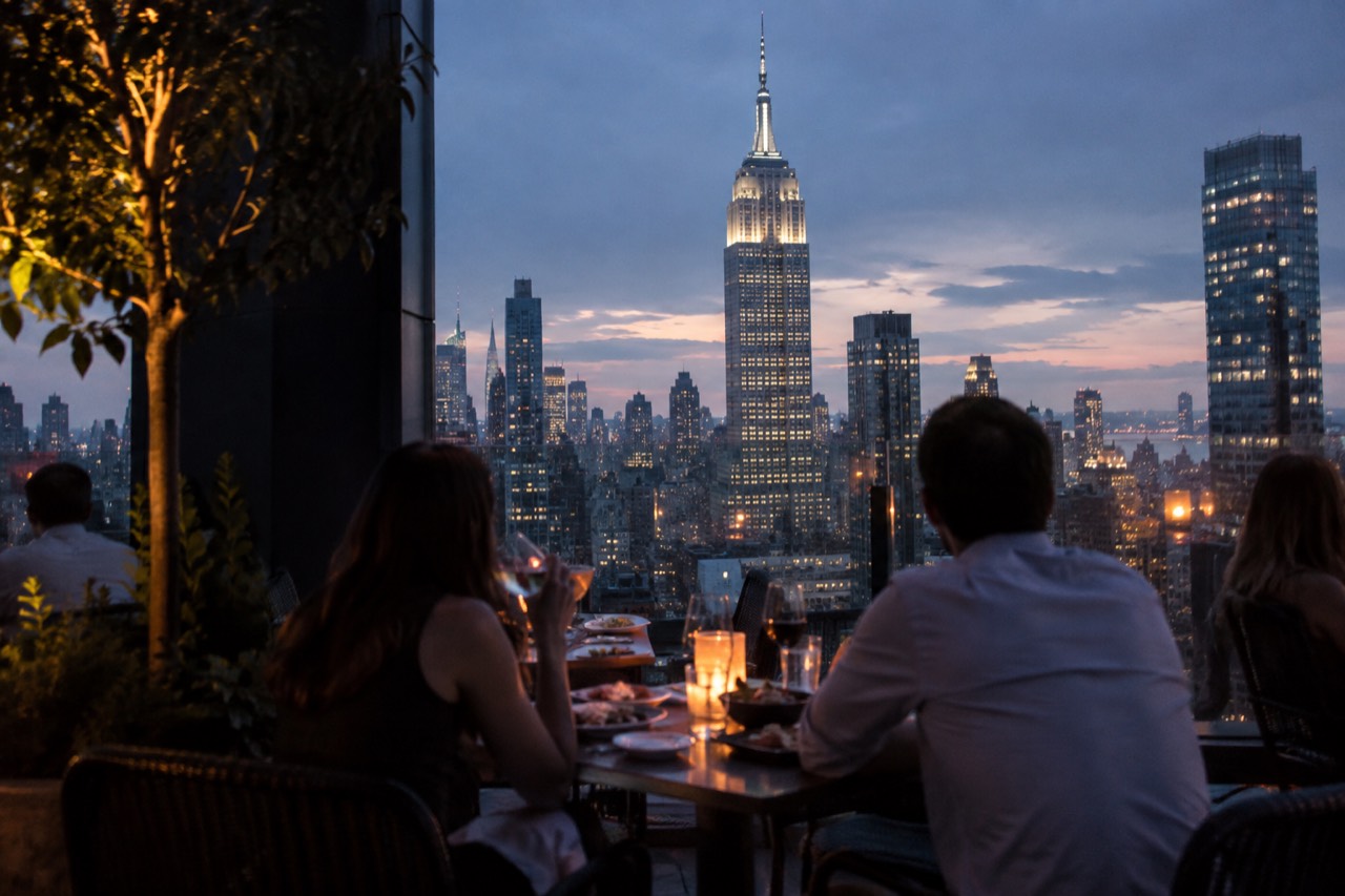 Diners at a rooftop restaurant table with the Empire State Building lit up in the evening skyline behind them