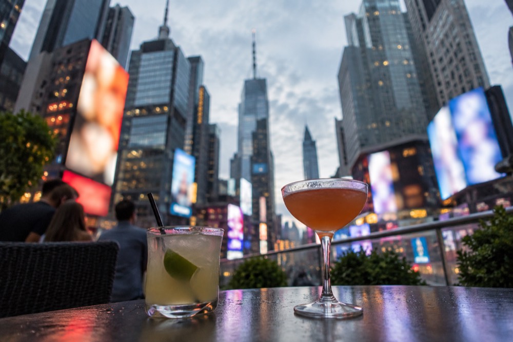 Two cocktails on a rooftop bar table with the illuminated signs of the Theater District in the background.