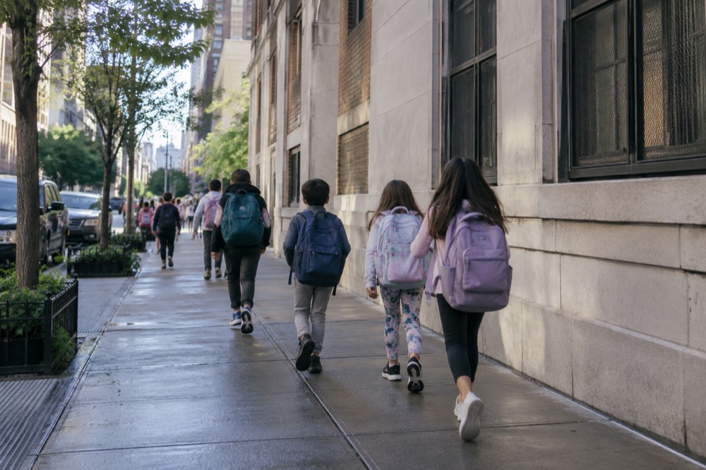 Children with backpacks walking past a New York City school building on a sunny morning