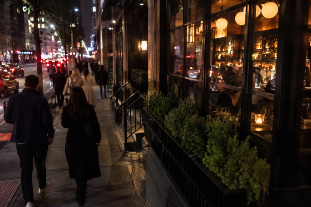 People walking past lit-up bar windows on a New York sidewalk at night