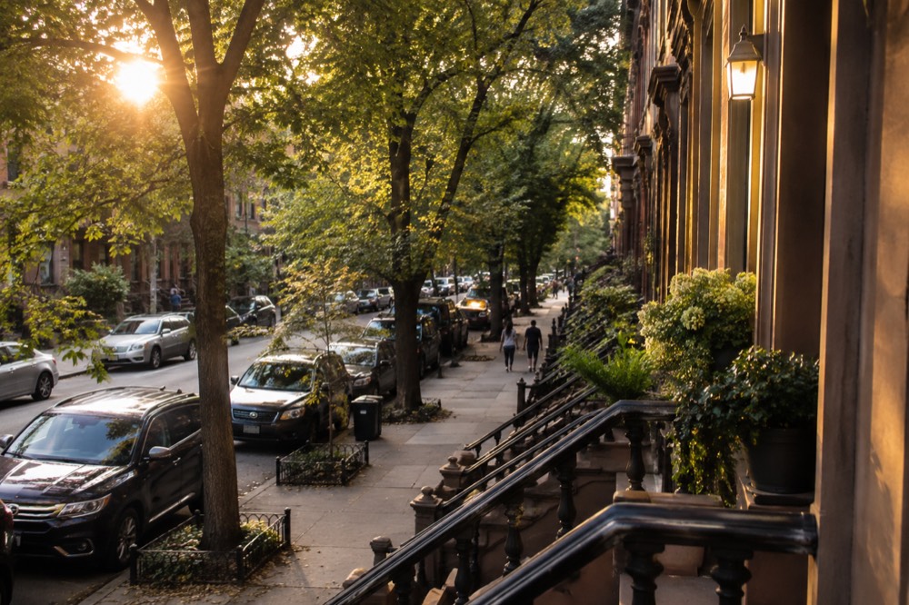 Cars parked along a tree-lined brownstone street in Brooklyn with a subway sign visible at the corner