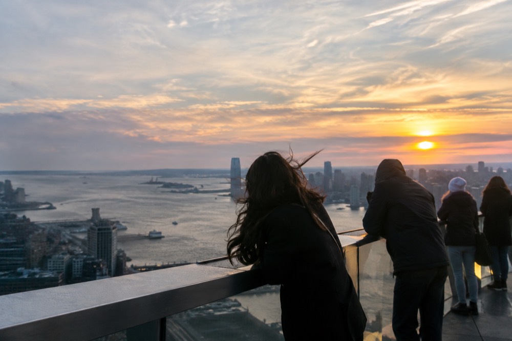 People at an outdoor railing wearing jackets with wind blowing as the sun sets over the river