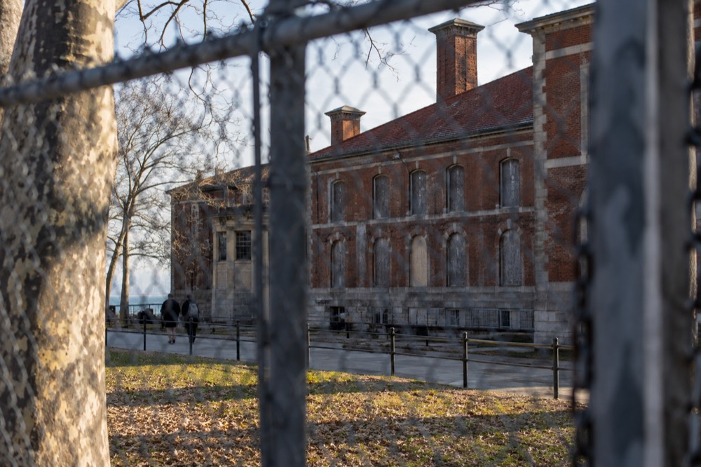 Exterior of the Ellis Island hospital complex with weathered brick walls and boarded windows seen across a fence