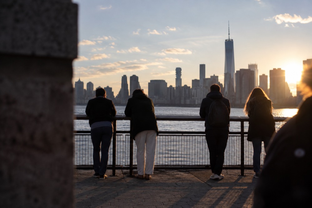 The Ellis Island promenade in late afternoon with visitors leaning on a railing looking toward Lower Manhattan