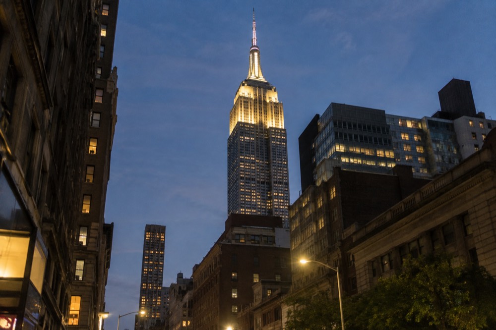 The Empire State Building lit at night, its floodlit tower visible above surrounding Midtown rooftops