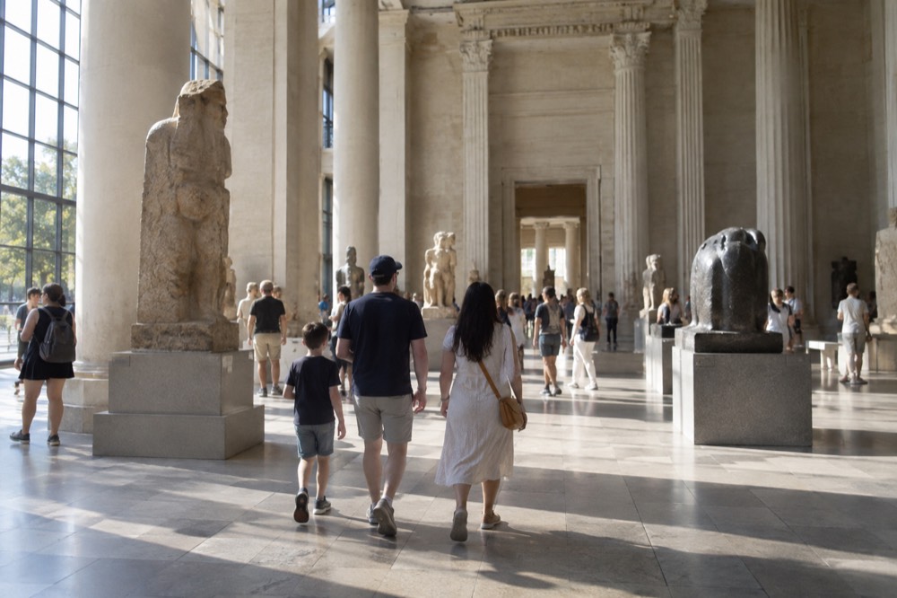 A family walking through a bright museum gallery with tall columns and stone artifacts