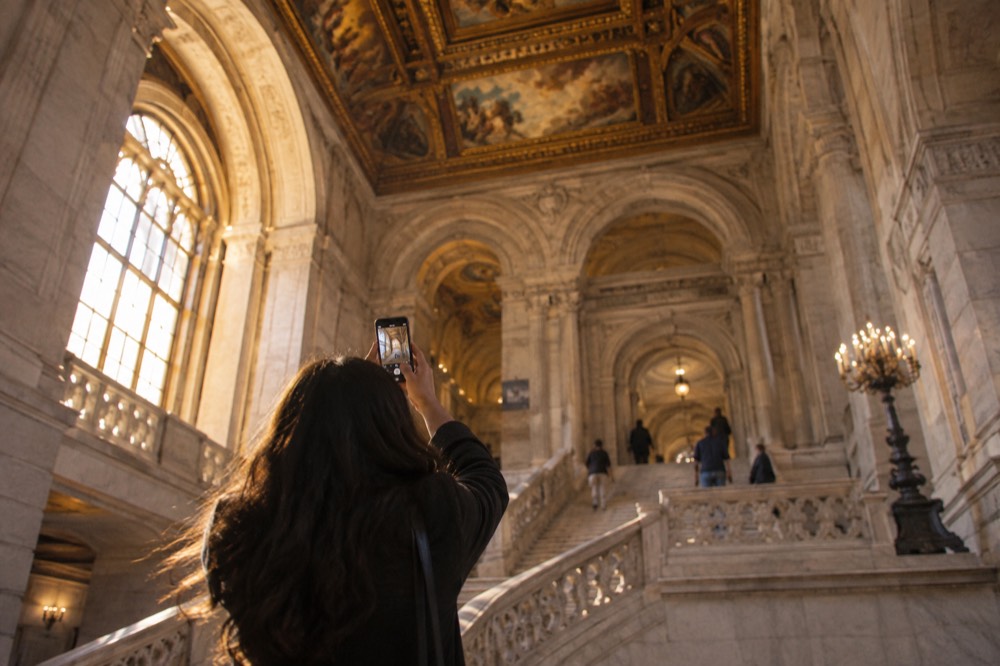 A visitor photographing the ornate painted ceiling above a marble staircase in the New York Public Library