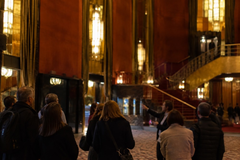 Visitors standing in a small group with a guide inside a ornate theater lobby with gold detailing
