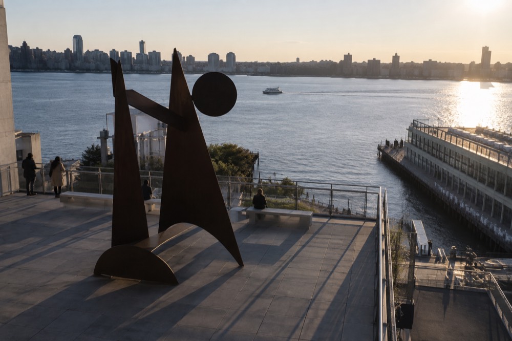 A large abstract sculpture displayed on an outdoor Whitney terrace with the Hudson River behind it