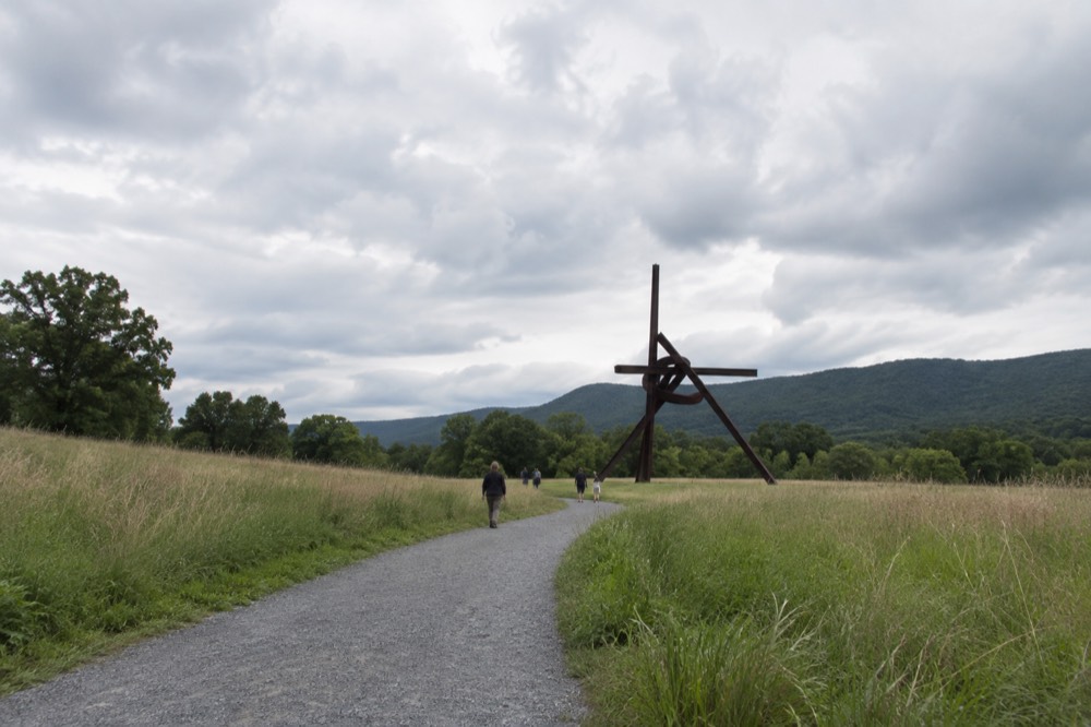 A wide gravel path curving through tall grass toward a tall steel sculpture