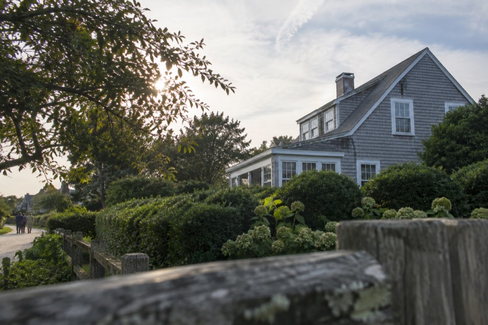 A weathered gray-shingled house behind hedges on a quiet Hamptons lane