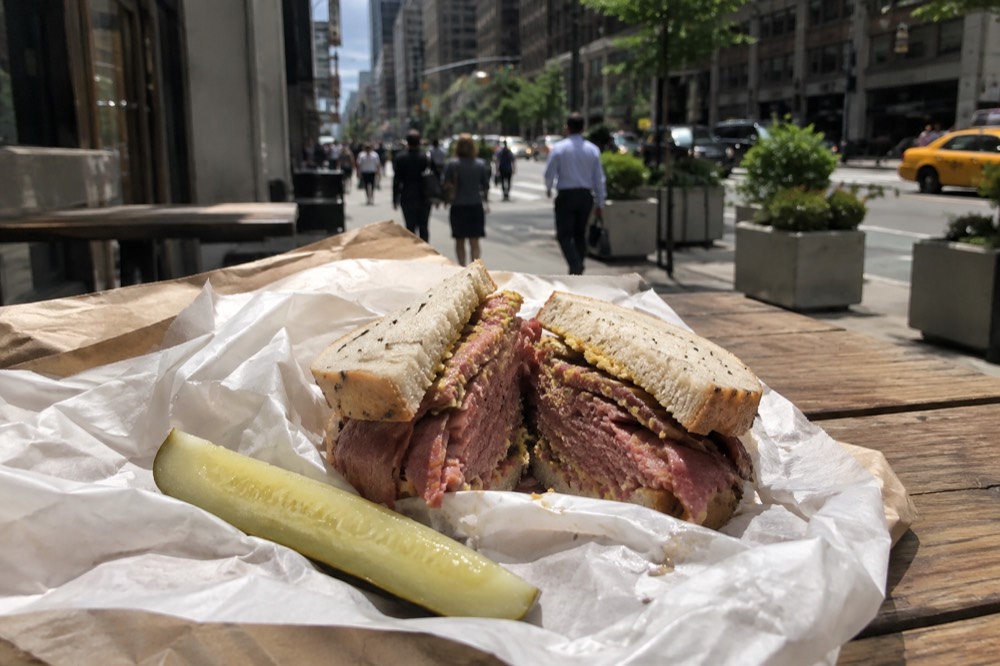 A sandwich wrapped in deli paper on a wooden table with a pickle spear