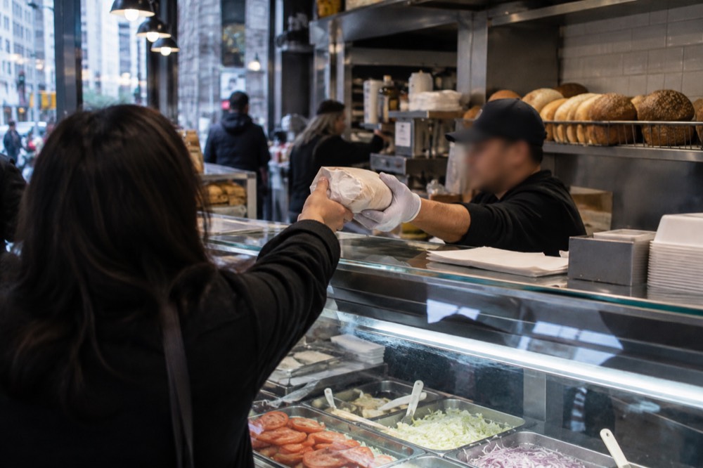 A counter worker handing a wrapped sandwich across a deli counter to a customer