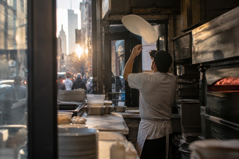 A cook tossing pizza dough in the air inside a small New York pizzeria kitchen
