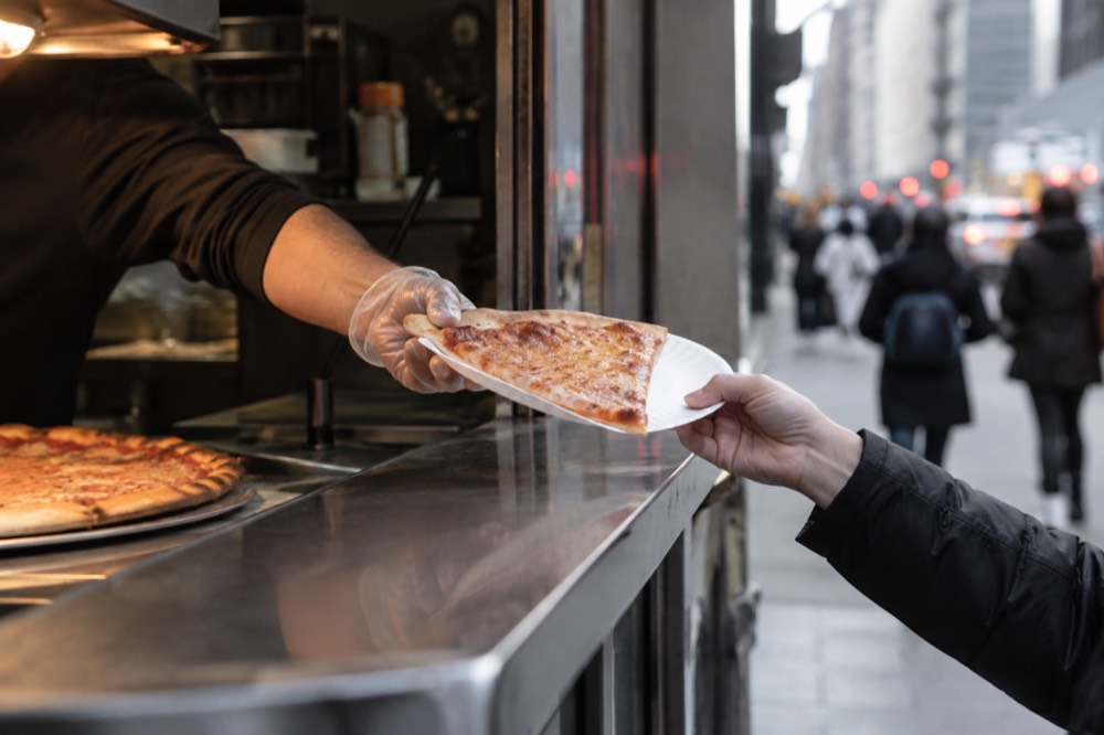 A cheese pizza slice being handed over a counter on a paper plate
