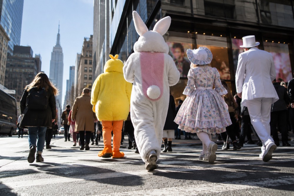 A costumed crowd crossing a New York street against a backdrop of lit storefronts