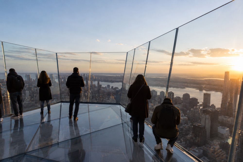 The glass floor and angled panels of an observation deck with visitors looking down at the city