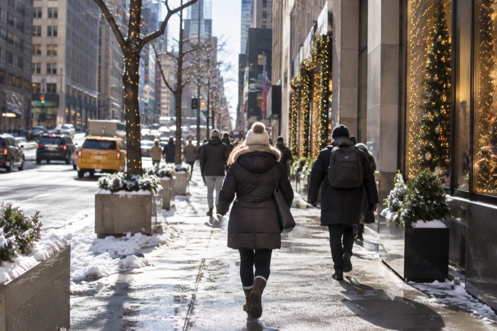 People in winter coats and hats walking a snow-dusted sidewalk past lit holiday windows