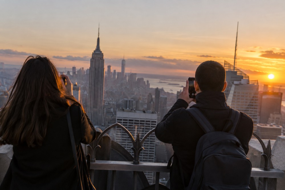 Two visitors photographing the Manhattan skyline at dusk from an outdoor observation deck