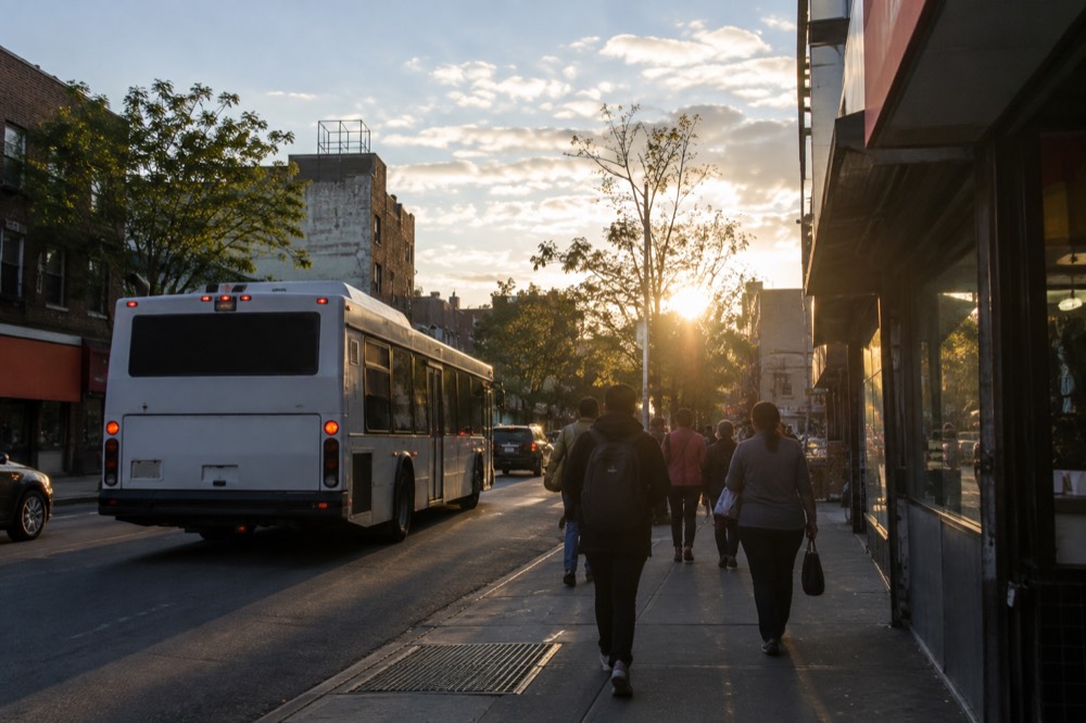 A city bus and walkers on a narrow street in Jackson Heights, Queens, lined with small shops
