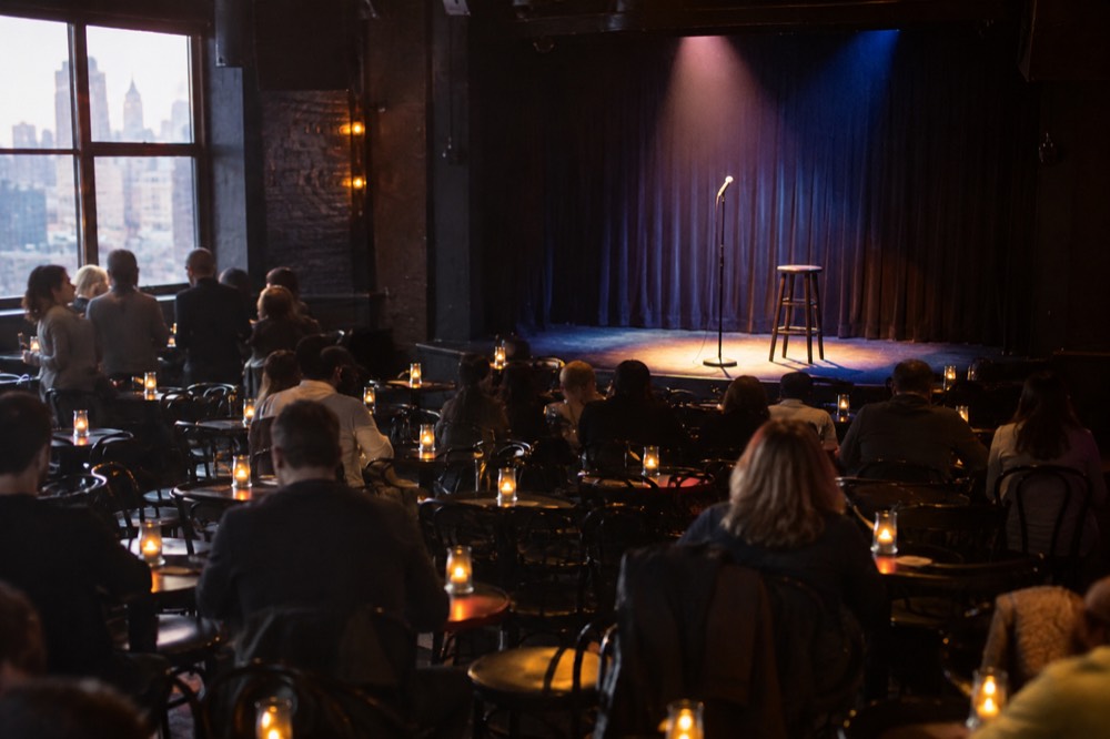 A small comedy club stage with a microphone stand under a spotlight and empty seats filling before a show