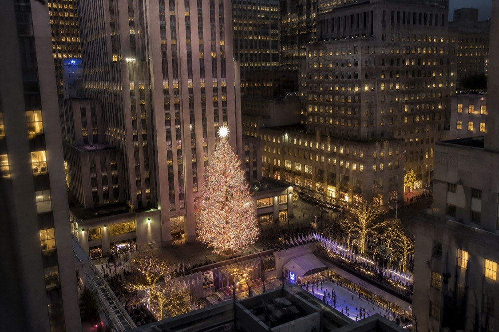 An illuminated Christmas tree seen from above, framed by city streets and lit buildings at night