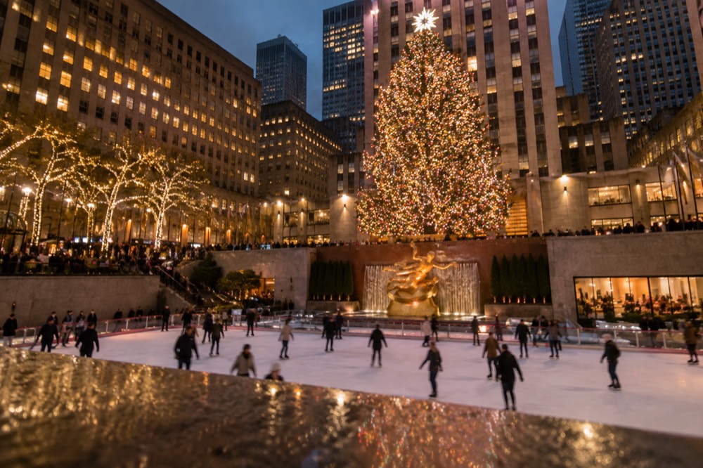 A lit Christmas tree glowing above an ice rink with skaters and tall buildings at night
