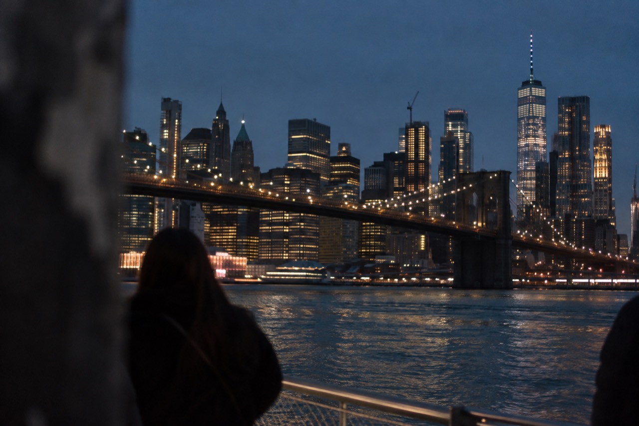 The Manhattan skyline and Brooklyn Bridge lit up against a dark night sky, seen from the waterfront