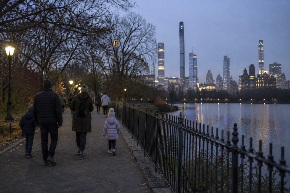 Families walking along a wide park path with a lake and city towers behind them