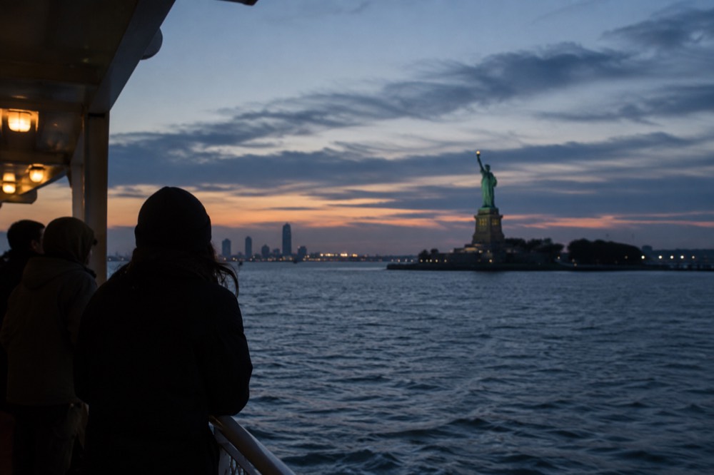 A ferry boat crossing a harbor toward a small island with a large statue