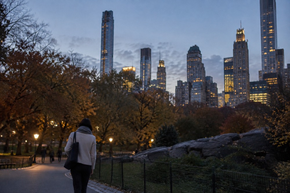 A person in a light jacket and scarf walking past autumn trees in a New York park