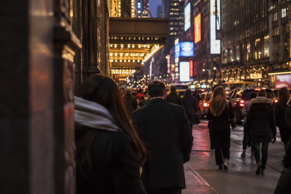 A busy Midtown sidewalk at night with theater marquees lit up along the block