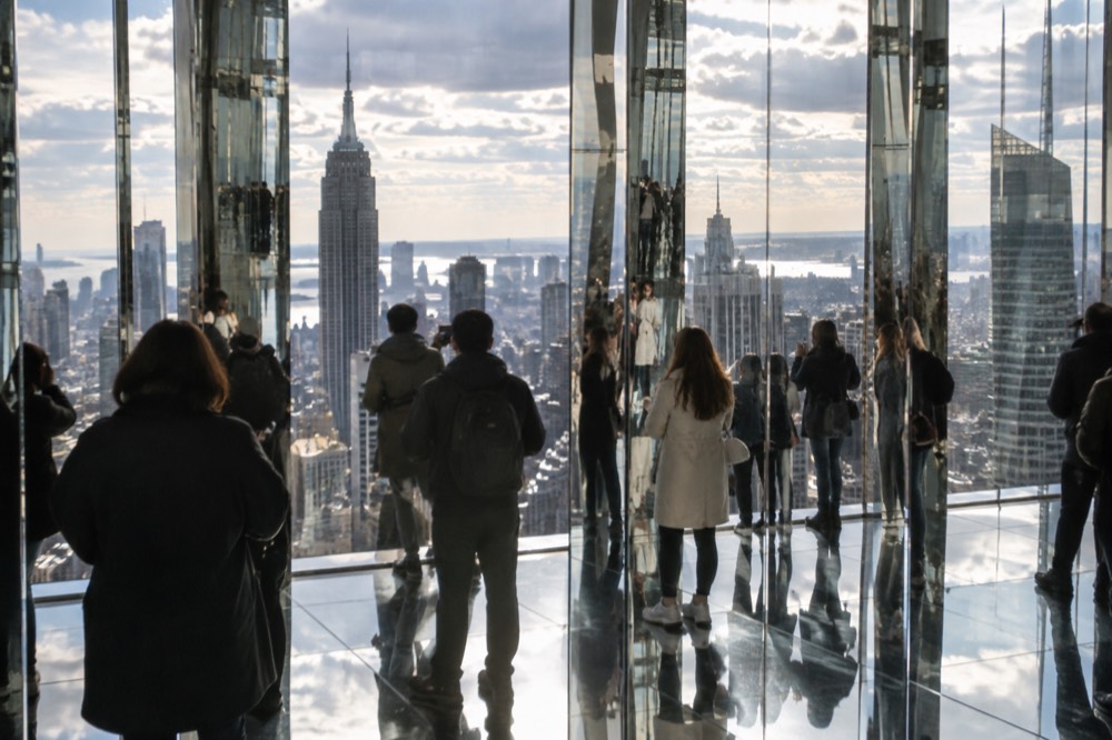 Visitors in a mirrored observation room high above Midtown Manhattan with reflections stretching across the glass